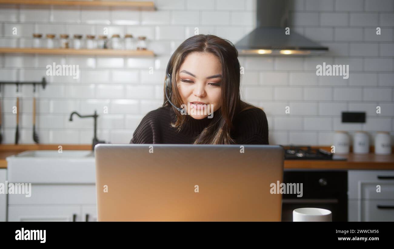 Asian woman wear wireless headset having video conference call on laptop computer talk by webcam in online chat in kitchen at home. Customer support s Stock Photo