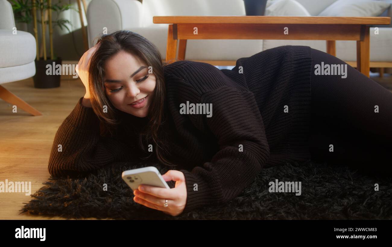 Smiling asian girl lying on floor surfing internet on mobile phone, checking email, reading media news, scrolling in social media, using mobile applic Stock Photo