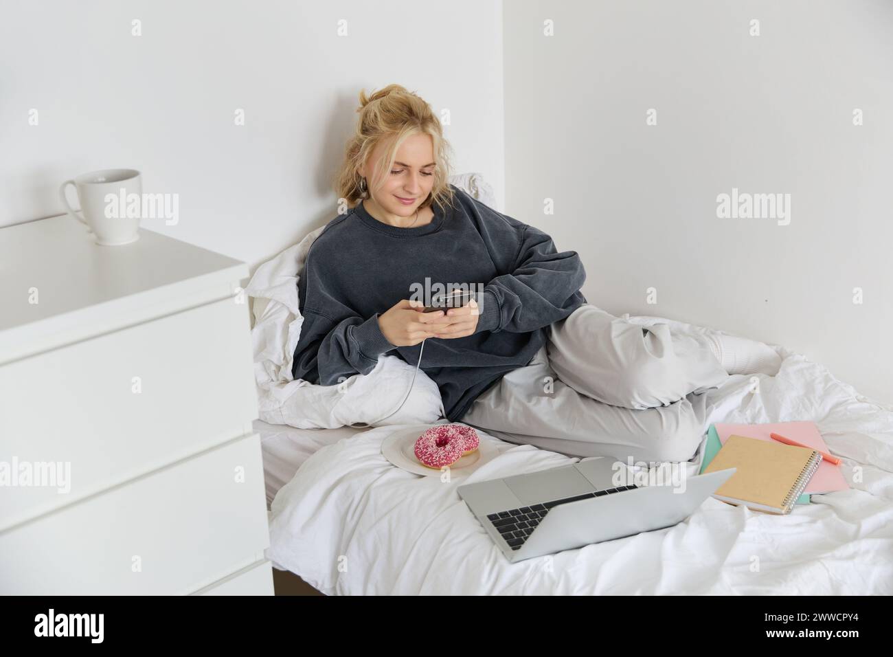 Portrait of smiling candid woman, lying in bed with doughnut, using smartphone and laptop, resting at home in bedroom, watching tv show or chatting on Stock Photo