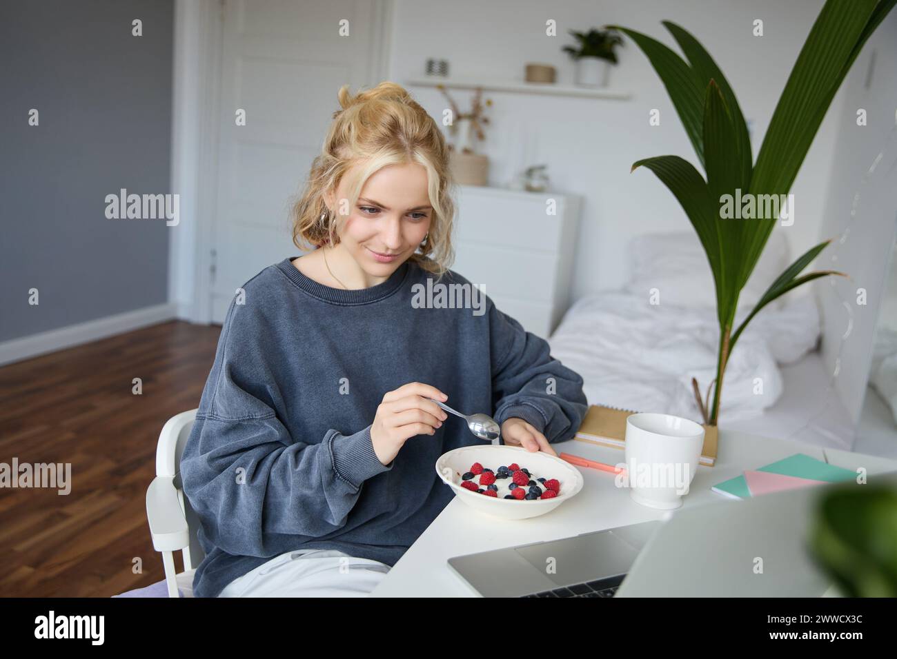 Portrait of young candid girl watching videos on laptop, enjoying movie and eating in front of laptop, having breakfast and staring at screen. Stock Photo