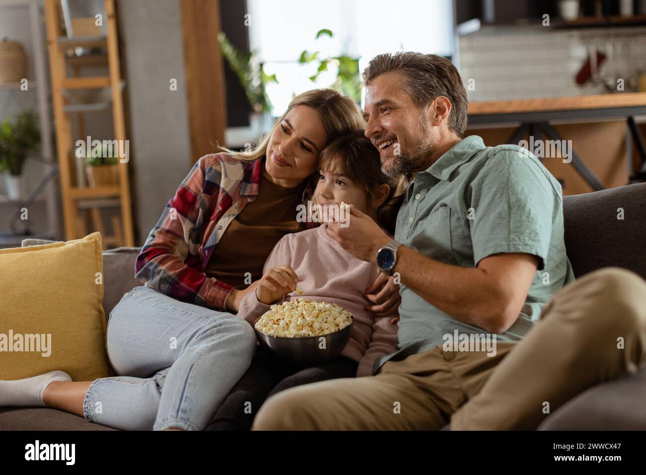 A family of three is comfortably nestled on a couch, their faces reflecting excitement and attentiveness as they share a bowl of popcorn during a susp Stock Photo