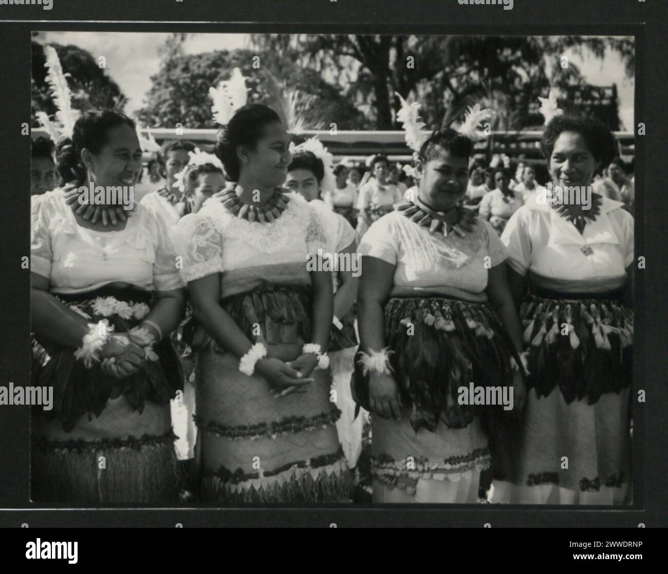 Description: Coy Tongan women dressed for dancing the Iskalaka await their turn to perform on the mala'e. They wear tauvala's of the fine woven mate adorned with scarlet parekeet feathers over the top of which are girdles of dracsens leaves. Location: Tonga tonga, australasia, oceania, australasiathroughalens Stock Photo