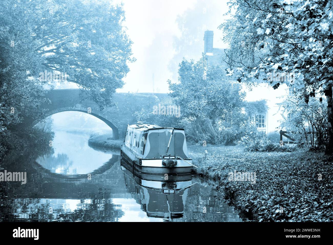 Atmospheric black & white image of a moored narrowboat near an arched bridge and canalside house on the Coventry Canal, Fradley, England, UK, Britain Stock Photo