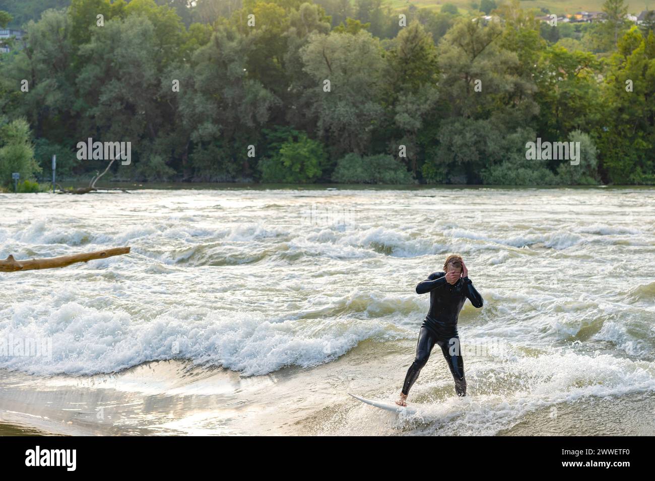 During the flood, a young surfer with neoprene wetsuit, is seen by the Aare river, wiping his face after braving the abundance of water. Stock Photo