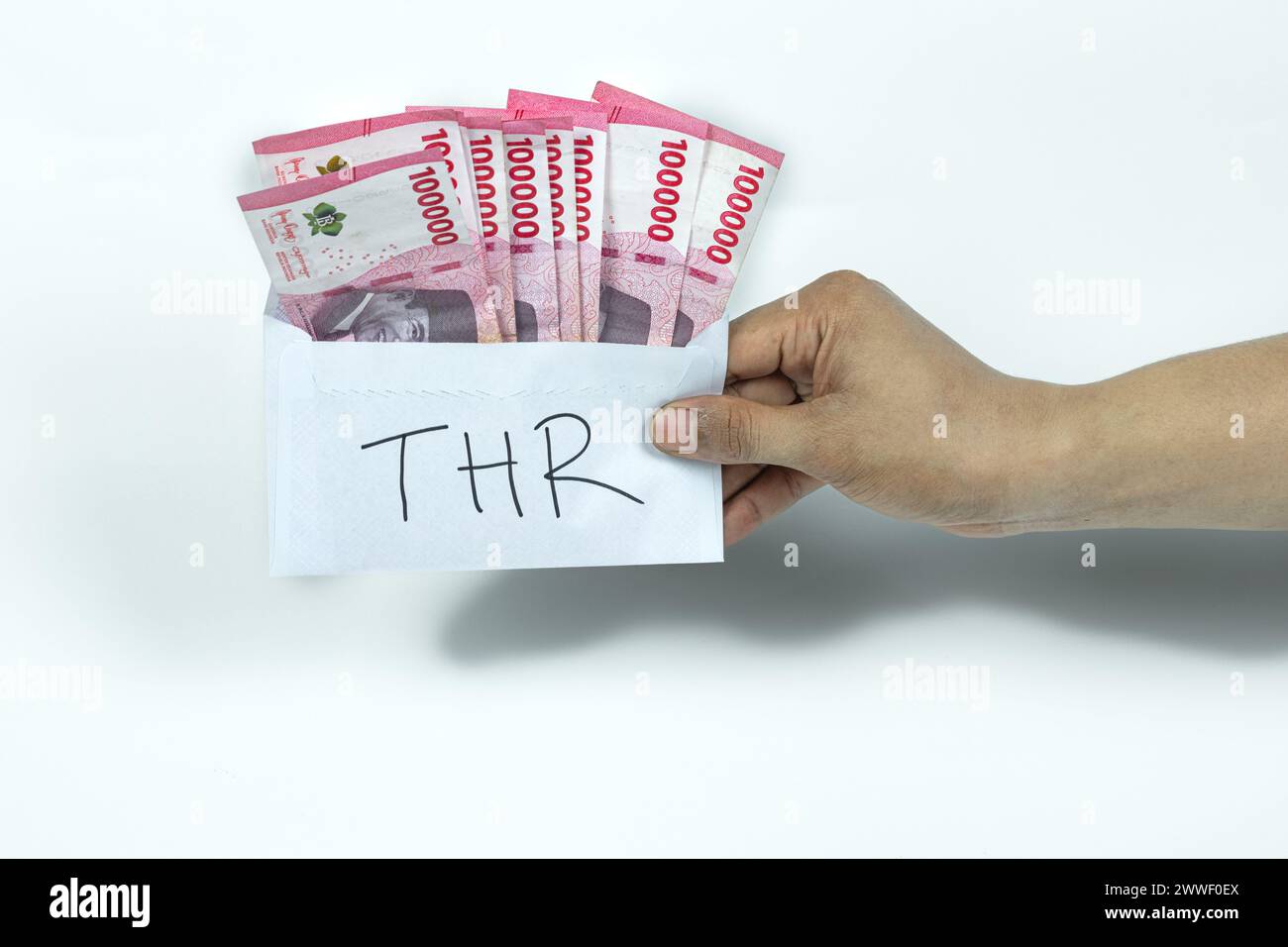 Asian man's hand holding white envelope with THR Text containing stack of IDR 100,000 cash.THR is holiday allowance on Eid al-Fitr or Lebaran days. Stock Photo