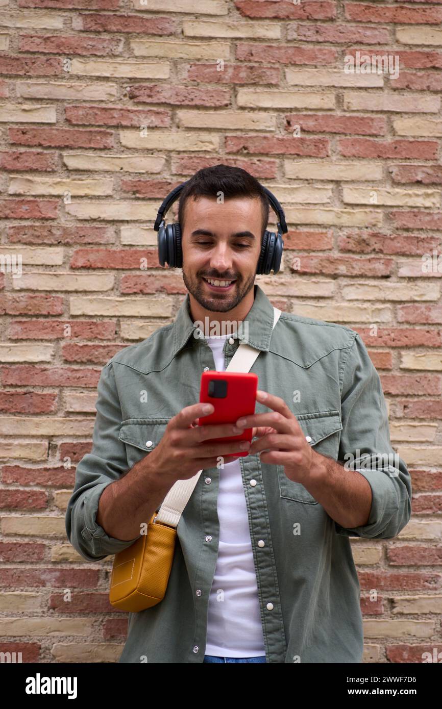 Handsome millennial man using mobile leaning against brick wall. People addicted technology.  Stock Photo