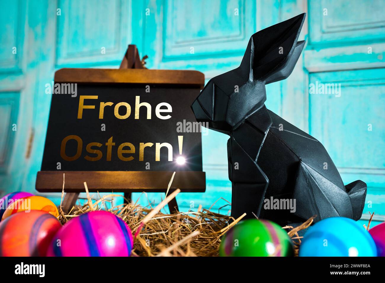 Augsburg, Bavaria, Germany - 23 March 2024: Easter nest with colorful Easter eggs in straw in front of an Easter bunny with a plaque and the lettering in golden letters: Happy Easter PHOTOMONTAGE *** Osternest mit bunten Ostereiern in Stroh vor einem Osterhasen mit einer Tafel und dem Schriftzug in goldener Schrift: Frohe Ostern FOTOMONTAGE Stock Photo