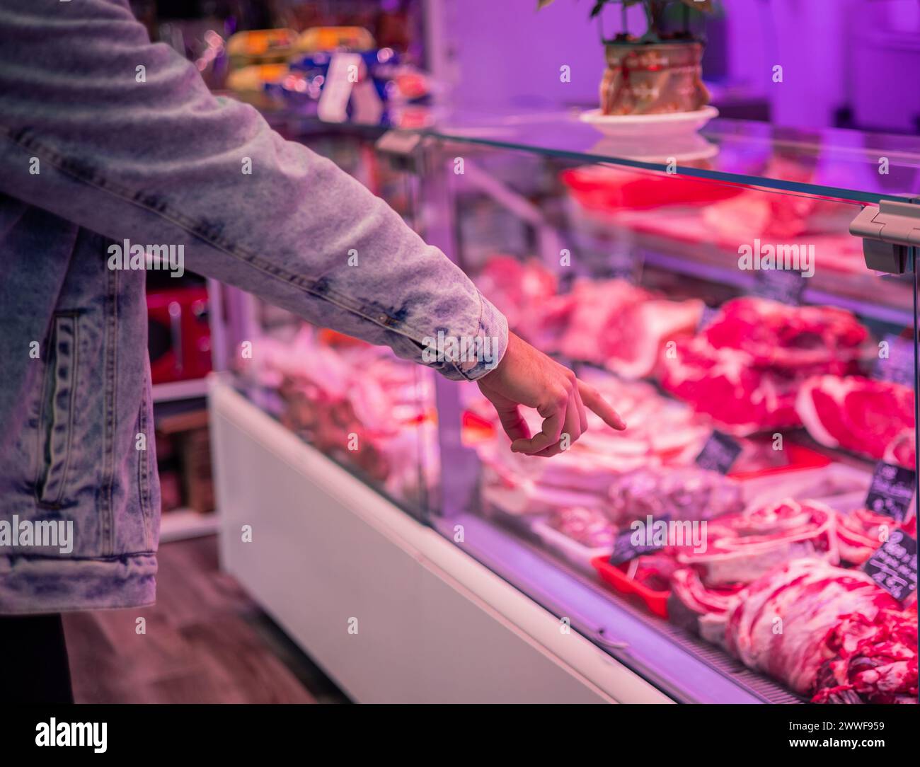 In this image, a customer is pointing with their finger at the products displayed on the counter of a butcher shop. Their hand and arm are in closeup, Stock Photo