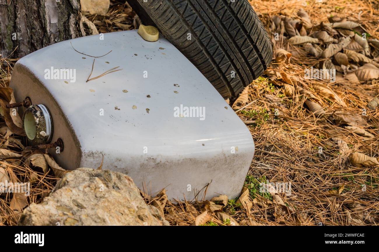 A white toilet tank resting on the ground near a tire surrounded by pine needles, in South Korea Stock Photo