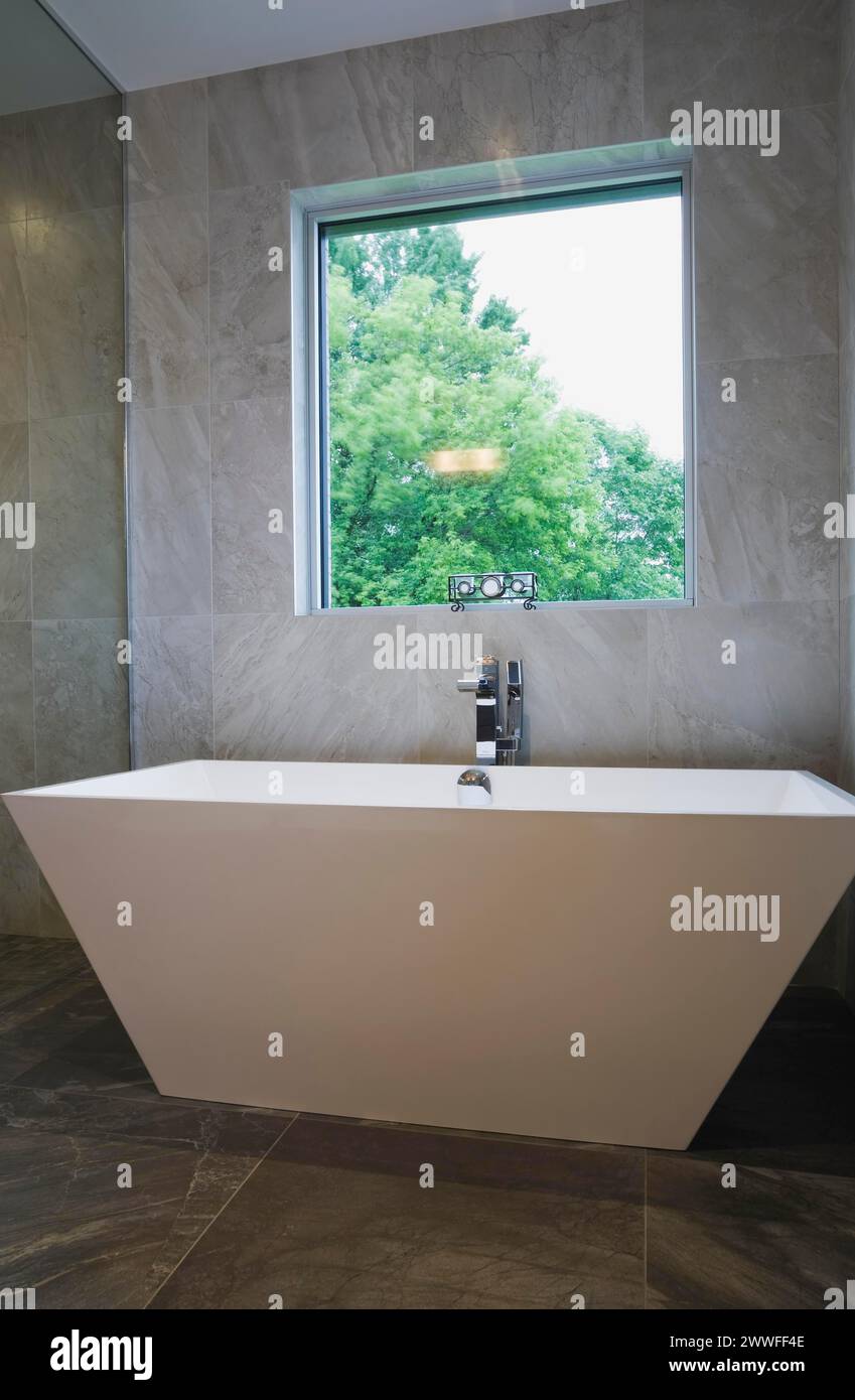 White freestanding vessel shaped bathtub in bathroom with grey ceramic tile floor and walls on ground floor inside modern cubist style home, Quebec Stock Photo