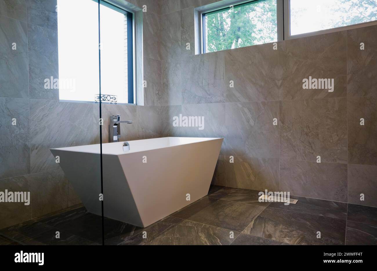 White freestanding vessel shaped bathtub in bathroom with grey ceramic tile floor and walls on ground floor inside modern cubist style home, Quebec Stock Photo