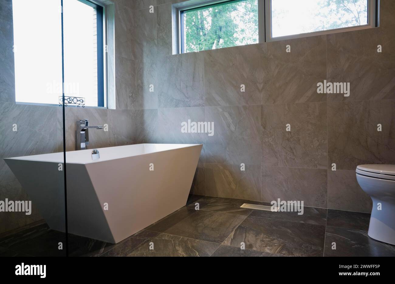 White freestanding vessel shaped bathtub in bathroom with grey ceramic tile floor and walls on ground floor inside modern cubist style home, Quebec Stock Photo