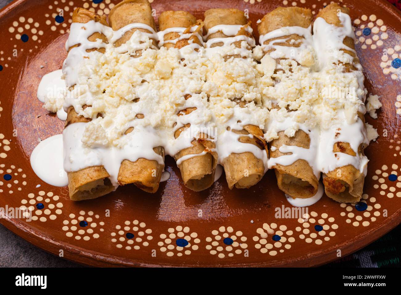 Fried potato tacos with cream and cheese in a Mexican mud dish. Tacos dorados, Mexican food. Stock Photo