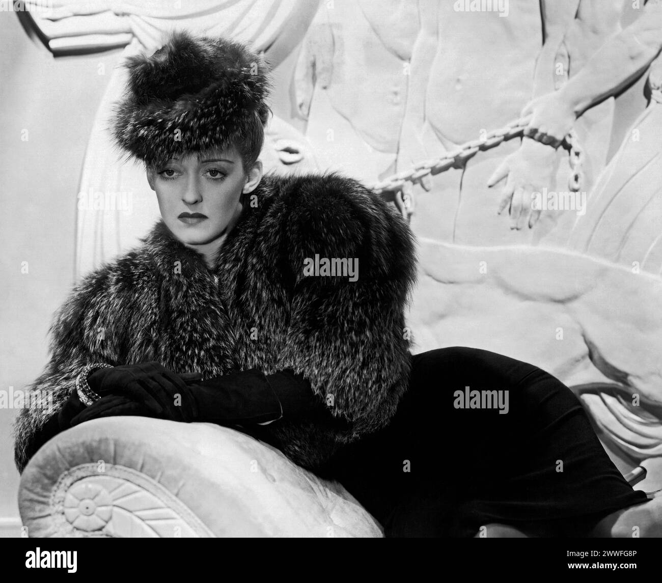 Hollywood, California   March 17, 1939 A studio portrait of actress Bette Davis wearing an elegant fur coat and matching hat while reclining on a stone couch with a sultry expression in those famous eyes. Stock Photo