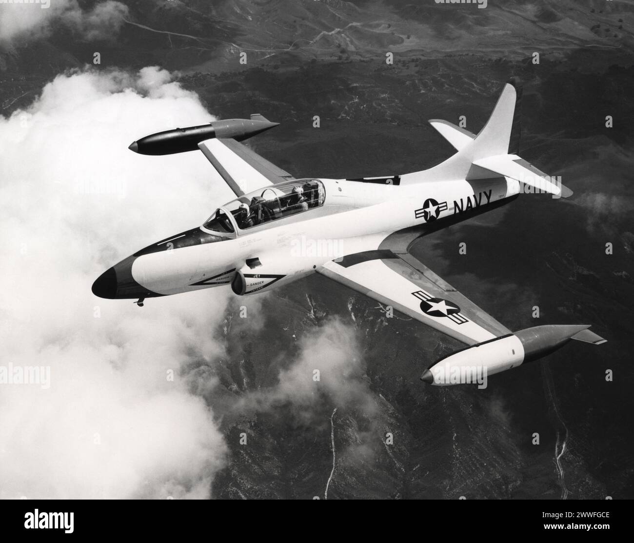 California, 1957 Silhouetted against the clouds so sharply it appears hand-painted by an artist, this T2V-1 SeaStar is just that. For Lockheed paint technicians, artists in their own field, have introduced a new skin-thin paint job on the U.S. Navy's new jet trainer. During a 24-hour beautifying session in Lockheed's paint shop, the SeaStar receives a four layer 'system' of paint consisting of a wash primer, regular primer and two coats of lacquer. By applying each successive coat while the previous one is still soft, all in a 24-hour period, better adhesion and durability are achieved, compan Stock Photo