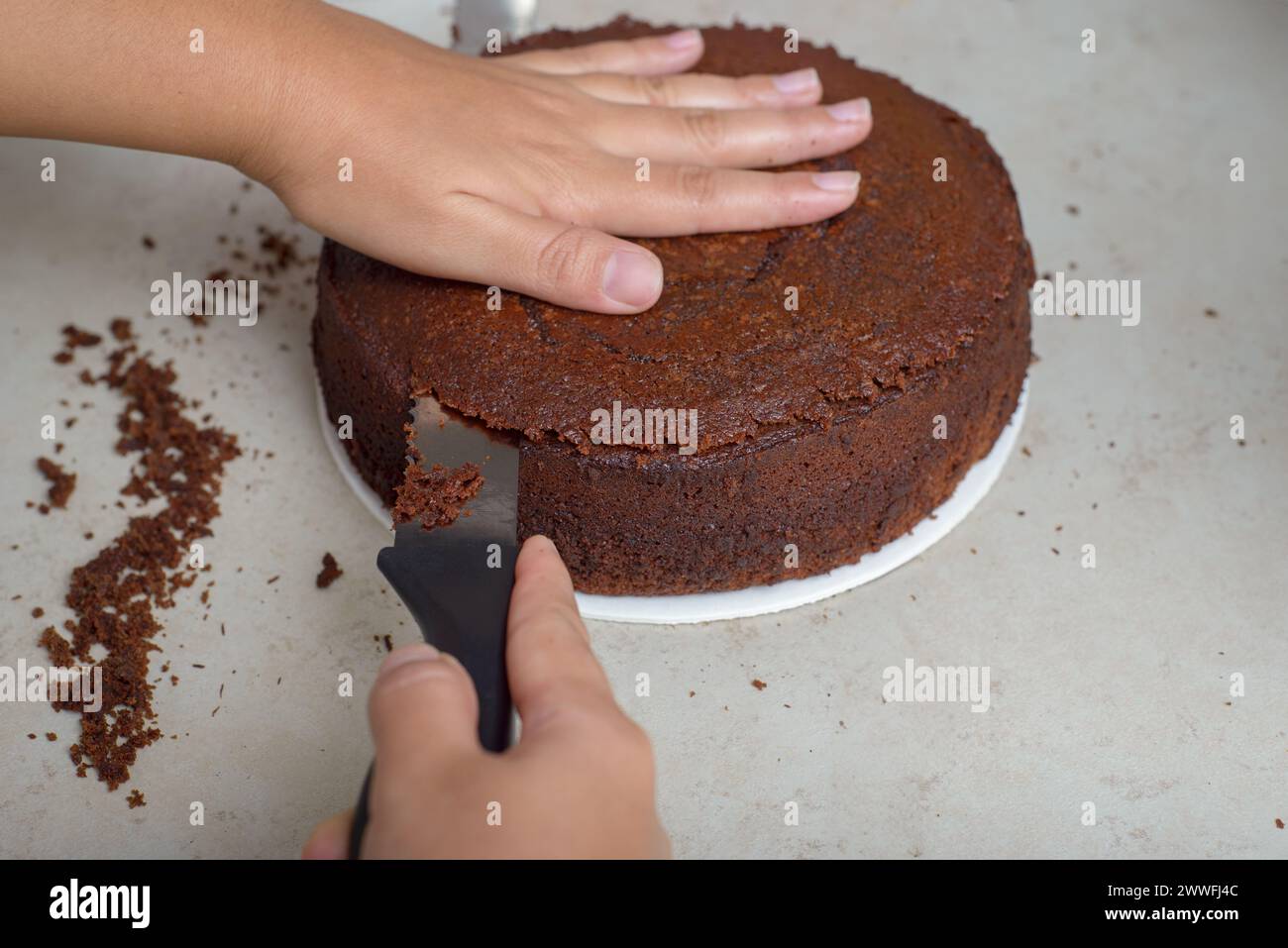 Person leveling a cake with a knife. Undecorated chocolate cake on wooden table. Stock Photo