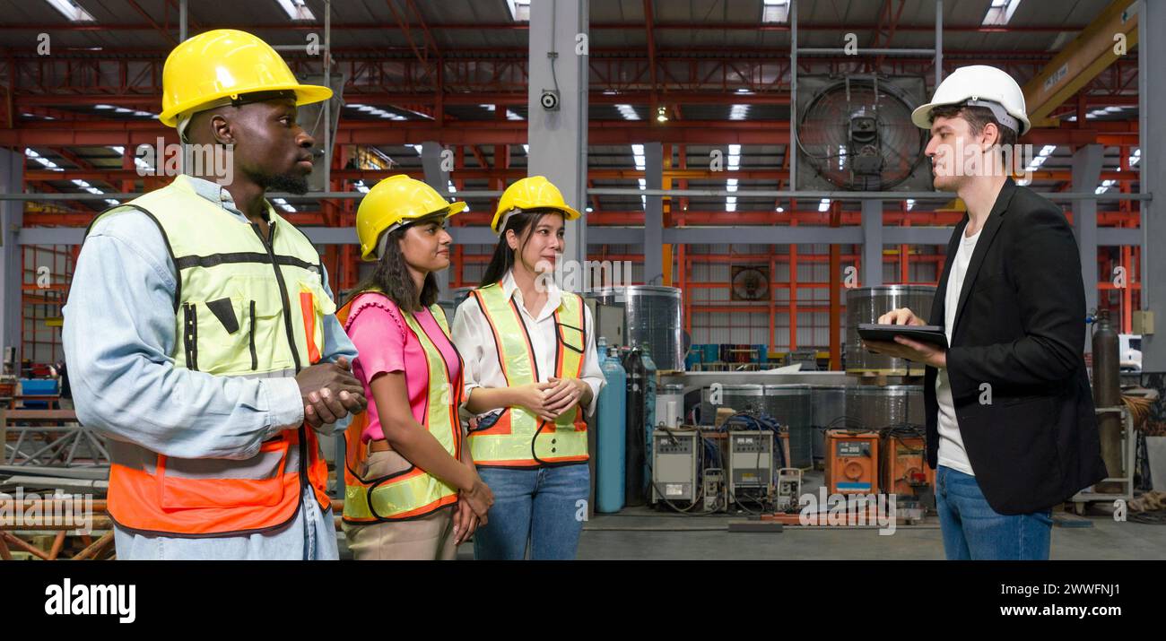 Group of engineer in safety vest meeting with young manager in black suit in an industrial or factory setting. Everyone wearing protective hardhat. At Stock Photo