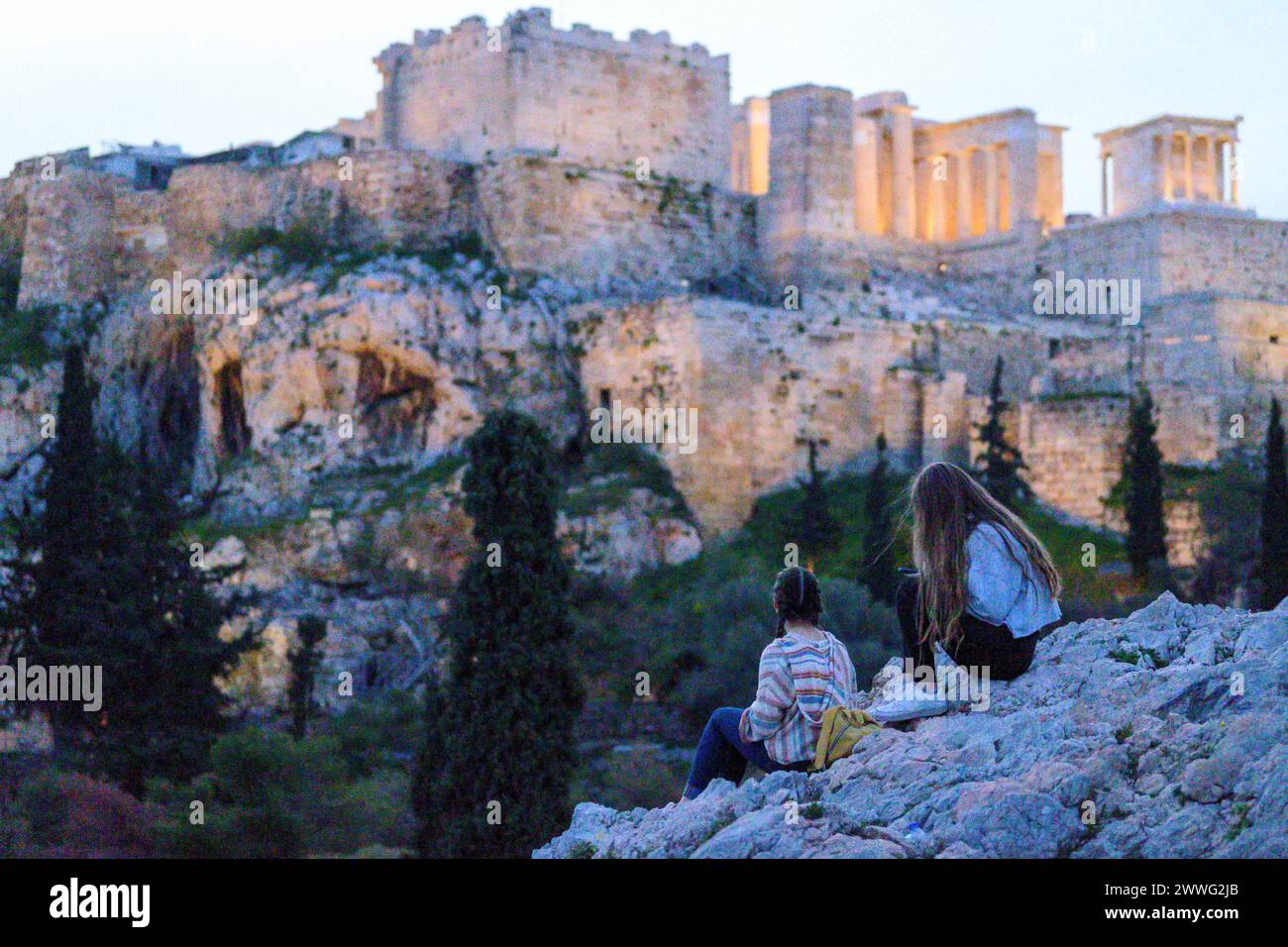 Two individuals seated on rocks, Acropolis in the background at sunset. Stock Photo