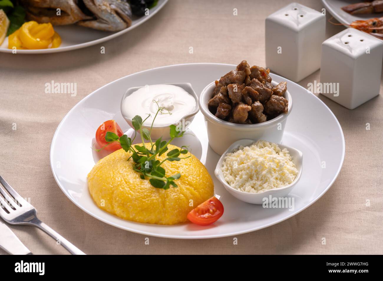Delicious polenta with cheese, stew meat and sour cream on white plate serving on table. Traditional romanian healthy food. Stock Photo