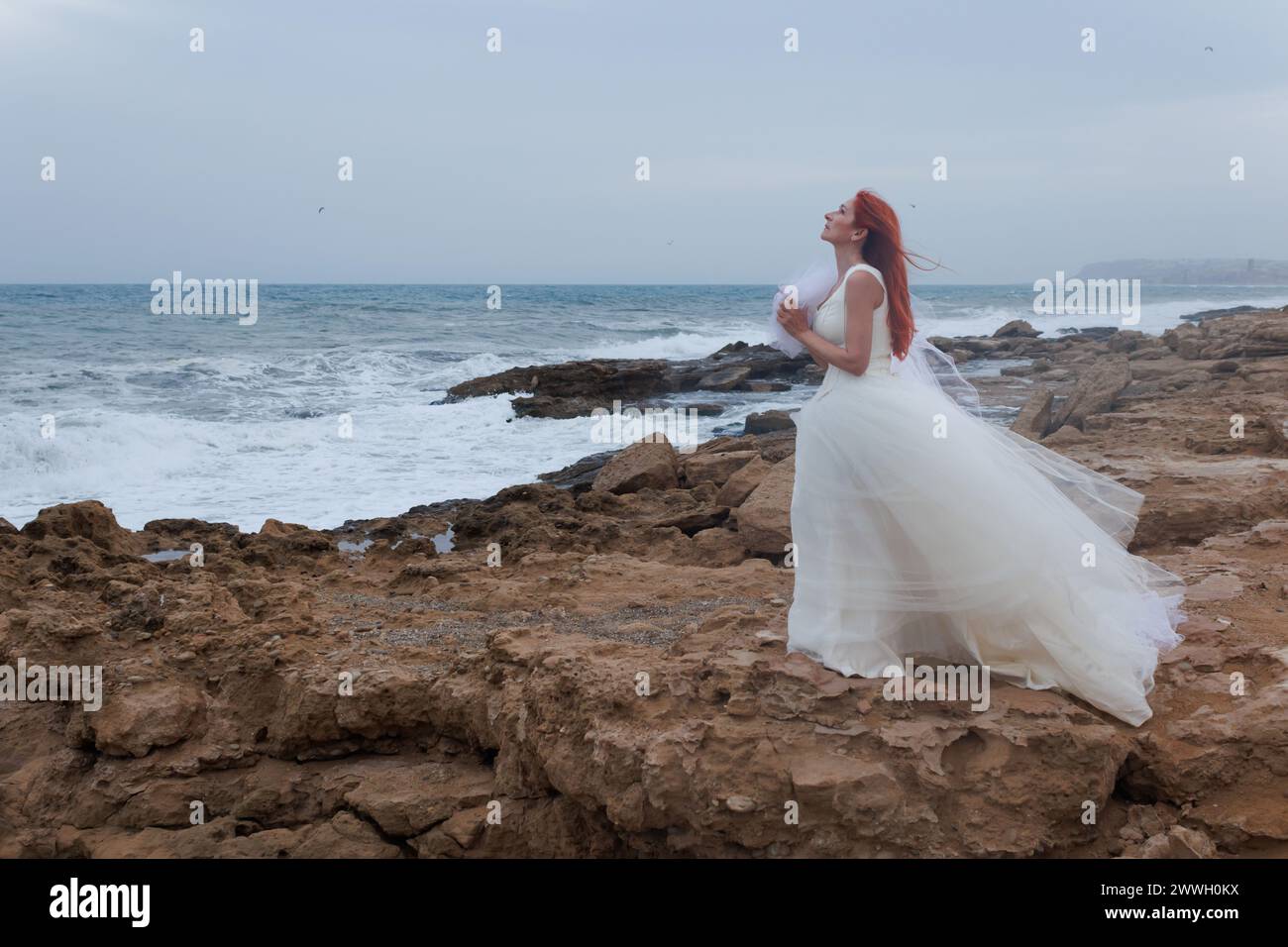 Lonely woman in wedding dress praying to gods a groom for wedding on windy day on Mediterranean sea coast, Alicante, Spain Stock Photo