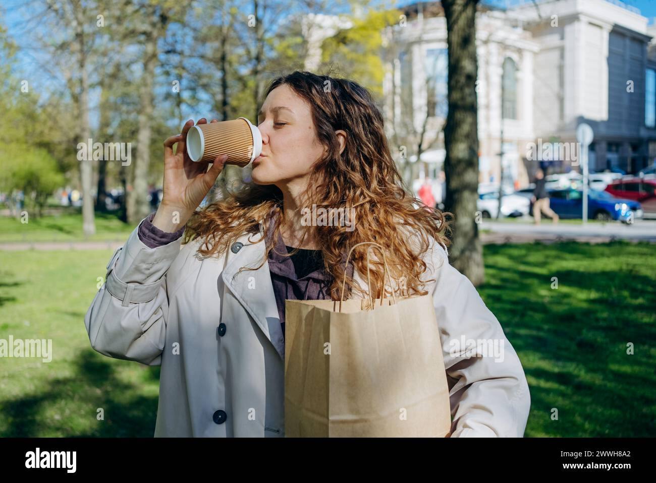 A woman savors her coffee from a takeaway cup while holding a shopping bag, relishing a sunny day in a vibrant city park. The woman bought lunch at a takeaway cafe in a craft paper bag. Stock Photo