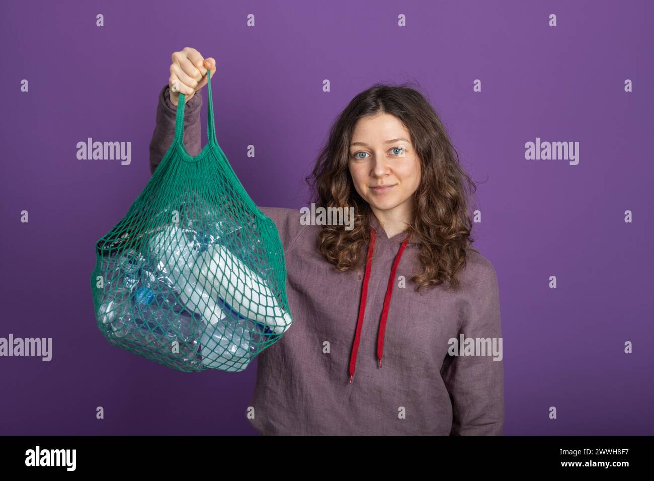 A woman with a gentle smile holds a green reusable mesh bag full of empty plastic bottles, promoting recycling and sustainability against a purple backdrop. Woman with Reusable Mesh Bag Stock Photo