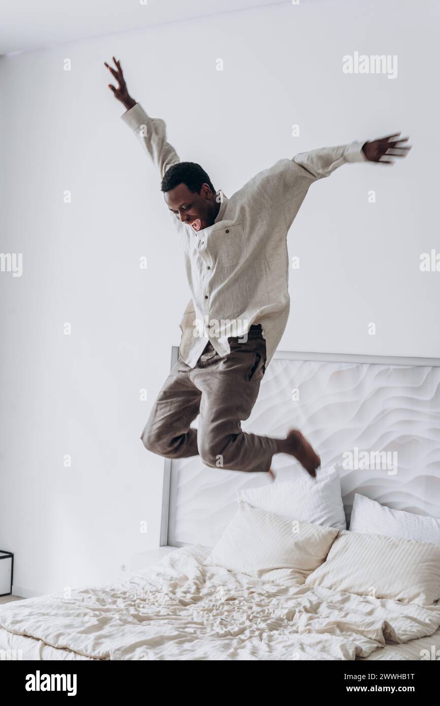 Exuberant joy radiates from a man as he leaps into the air above a bed, a spontaneous moment captured against a clean, white backdrop. Happy African American man jumps on the bed Stock Photo