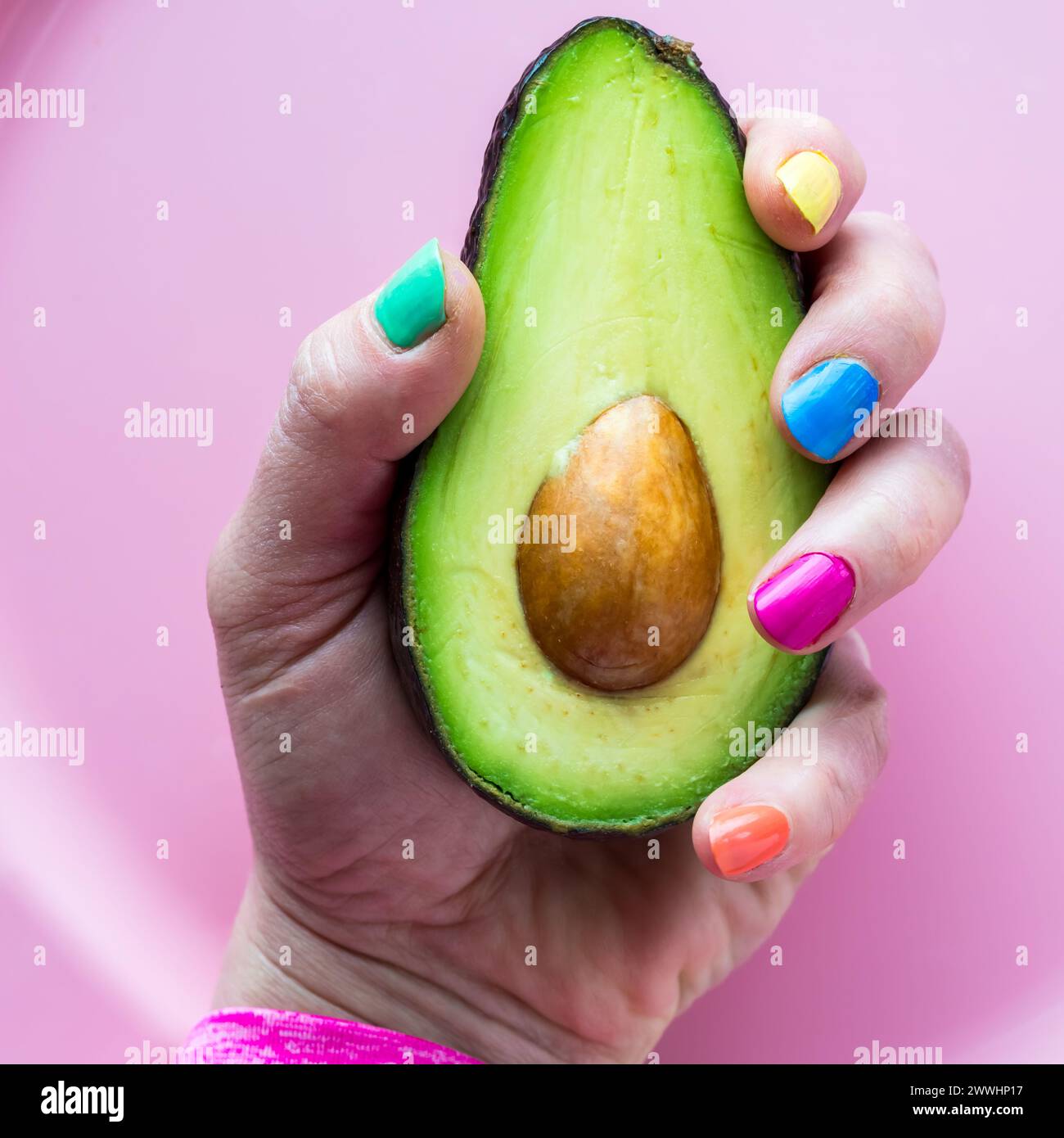 A hand with colourful nail polish holding a freshly cut avocado half. Stock Photo