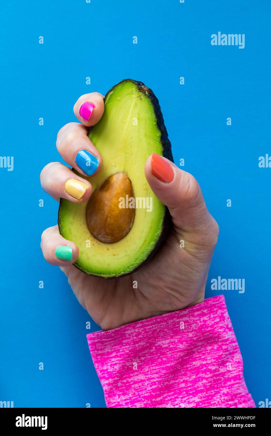 A hand with colourful nail polish holding a cut ripe avocado. Stock Photo