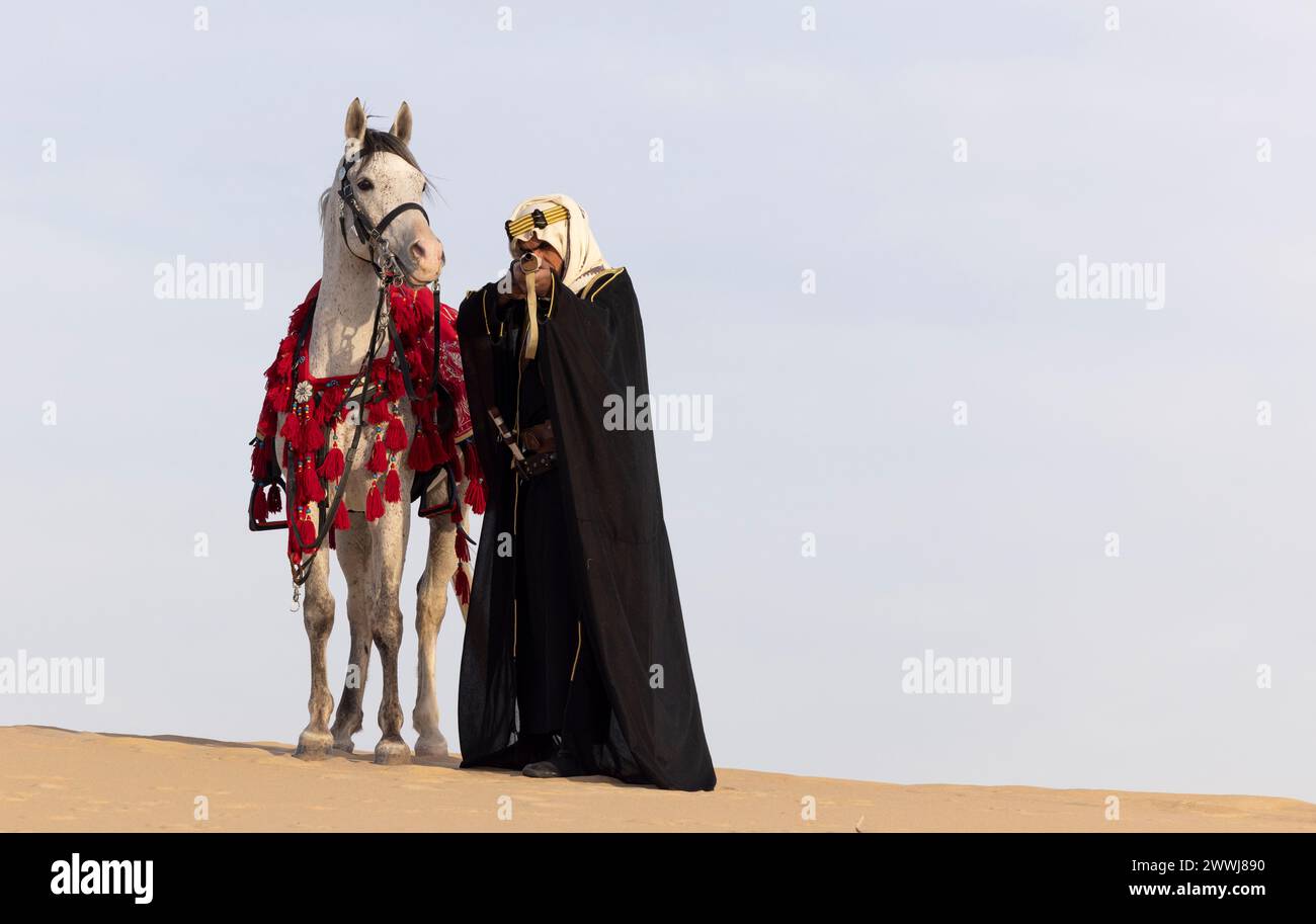 Saudi man in traditional clothing with his white stallion in a desert, aiming a rifle Stock Photo