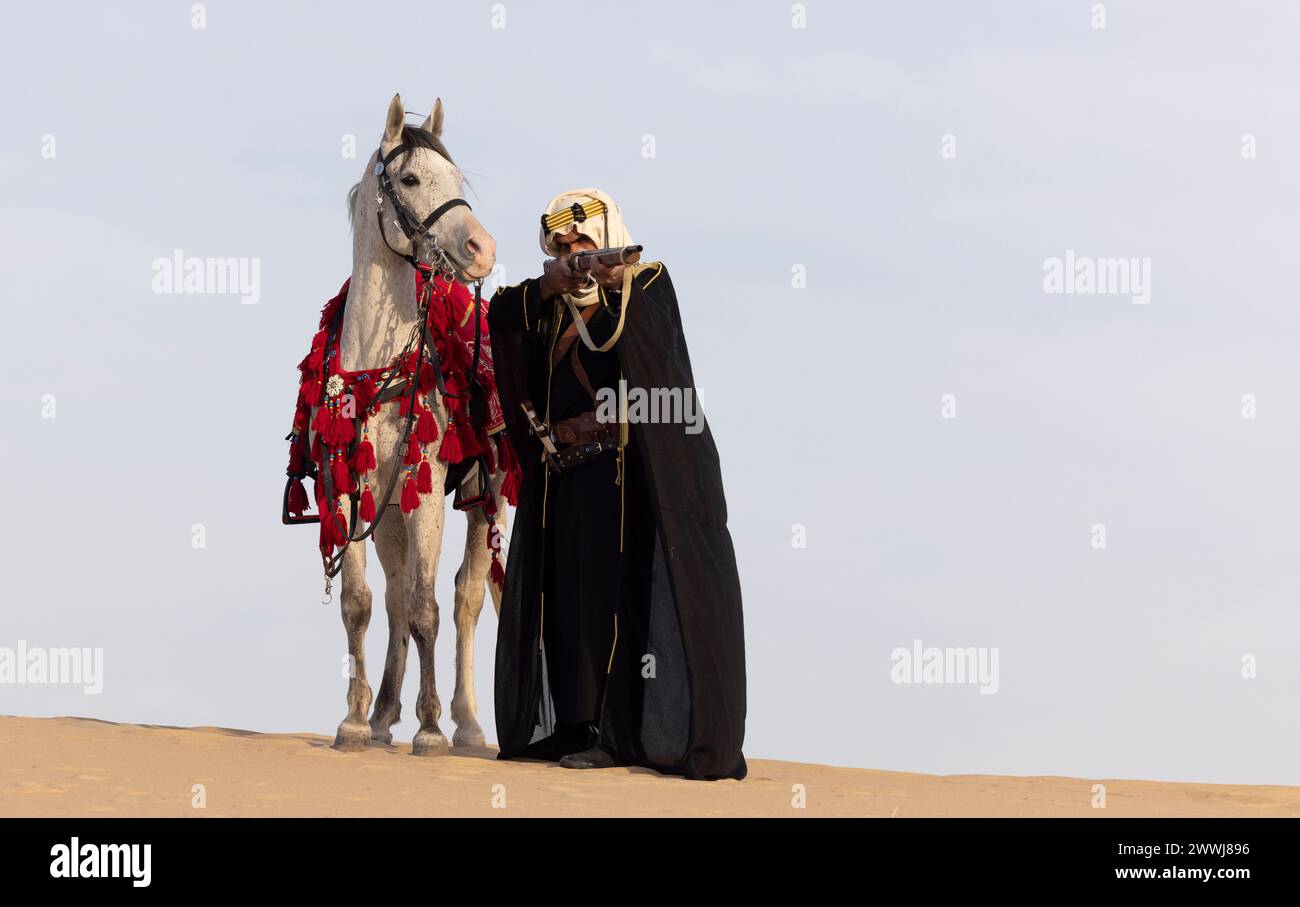 Saudi man in traditional clothing with his white stallion in a desert, aiming a rifle Stock Photo