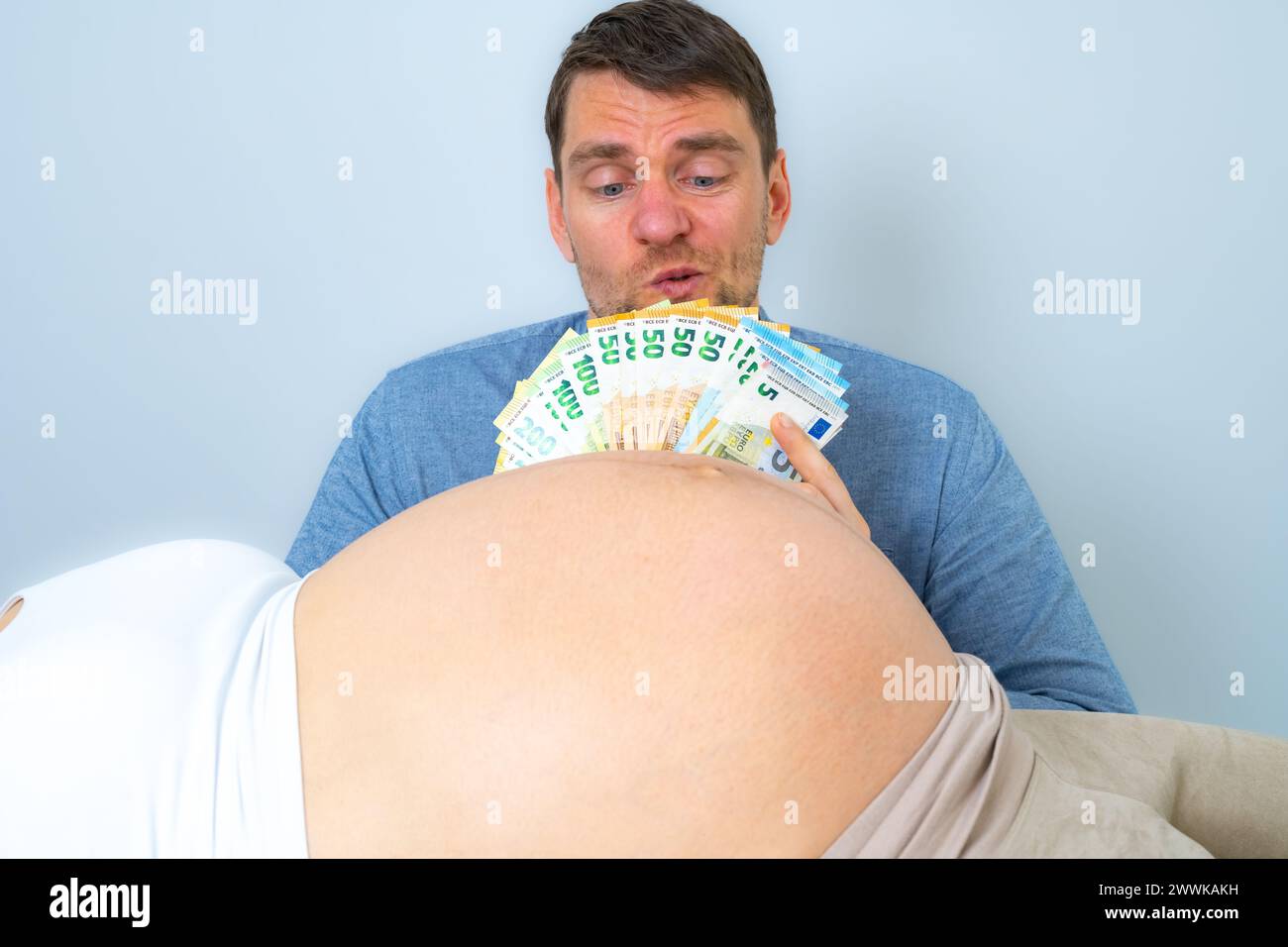 Description: Expectant father behind pregnant baby belly holds a stack of banknotes in front of his face and looks concerned. Last month of pregnancy Stock Photo