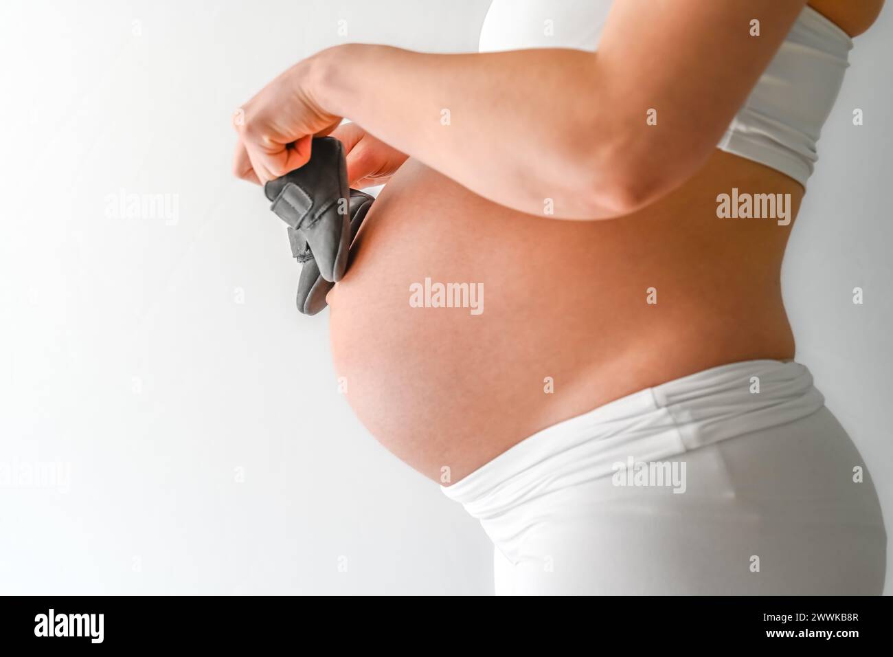 Description: Midsection of unrecognizable standing mother in white cloths walking baby shoes over her round pregnant belly. Final month of pregnancy - Stock Photo