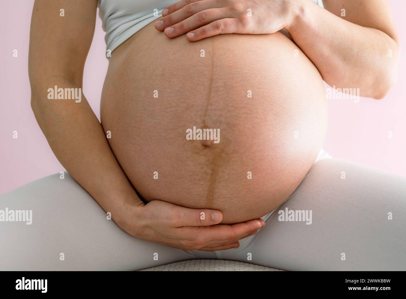 Description: Mother sits on a stool and gently holds her very round pregnant baby bump. Front view. Pink background. Bright shot. Stock Photo