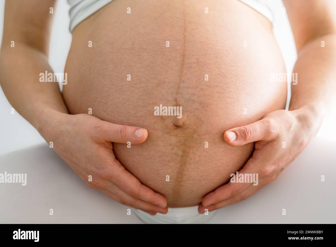 Description: Closeup of woman and gently holding her very round pregnant baby bump. Front view. White background. Bright shot. Stock Photo