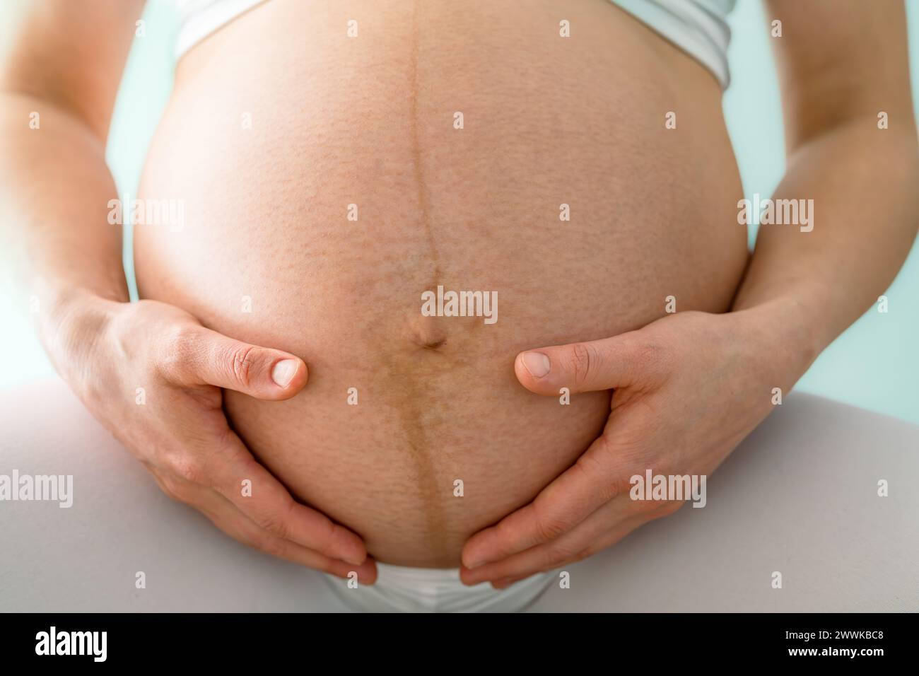 Description: Closeup of mother and gently holding her very round pregnant baby bump. Front view. Turquoise background. Bright shot. Stock Photo