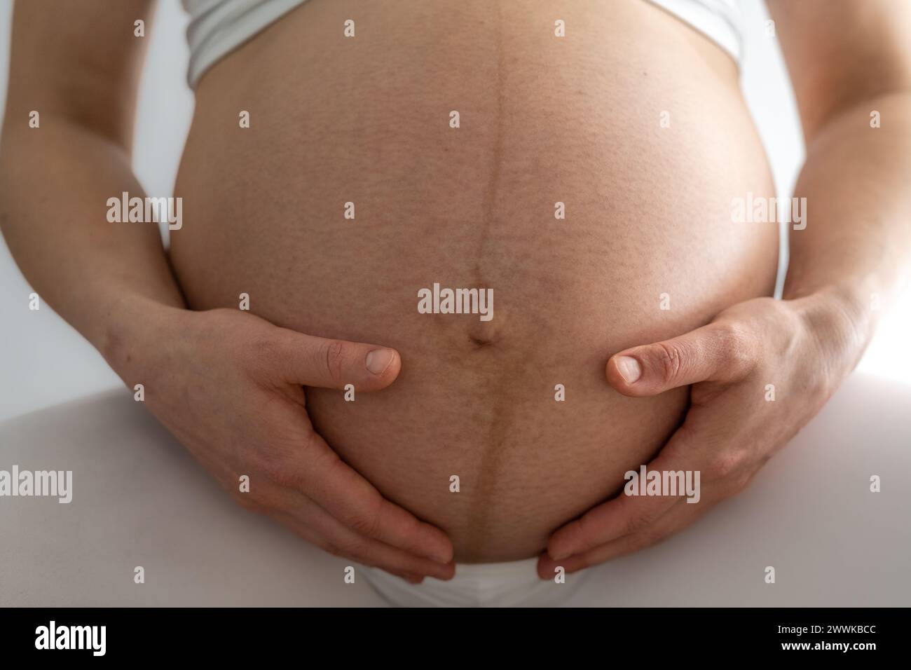 Description: Closeup of woman sitting and gently holding her very round pregnant baby belly. Front view. White background. Bright shot. Stock Photo