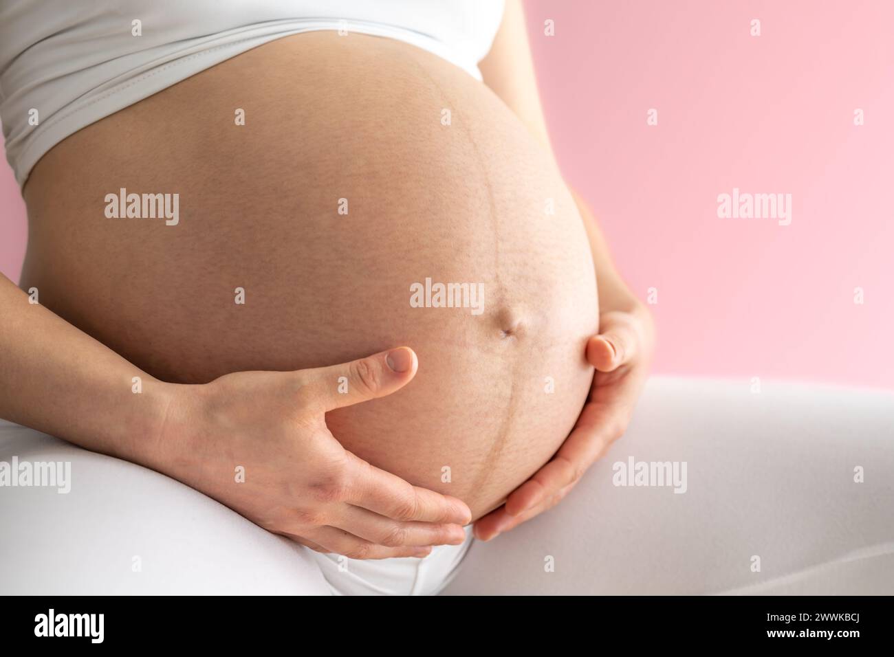 Description: Midsection of a mother and gently holding her very round pregnant baby bump. Side view. Pink background. Bright shot. Stock Photo