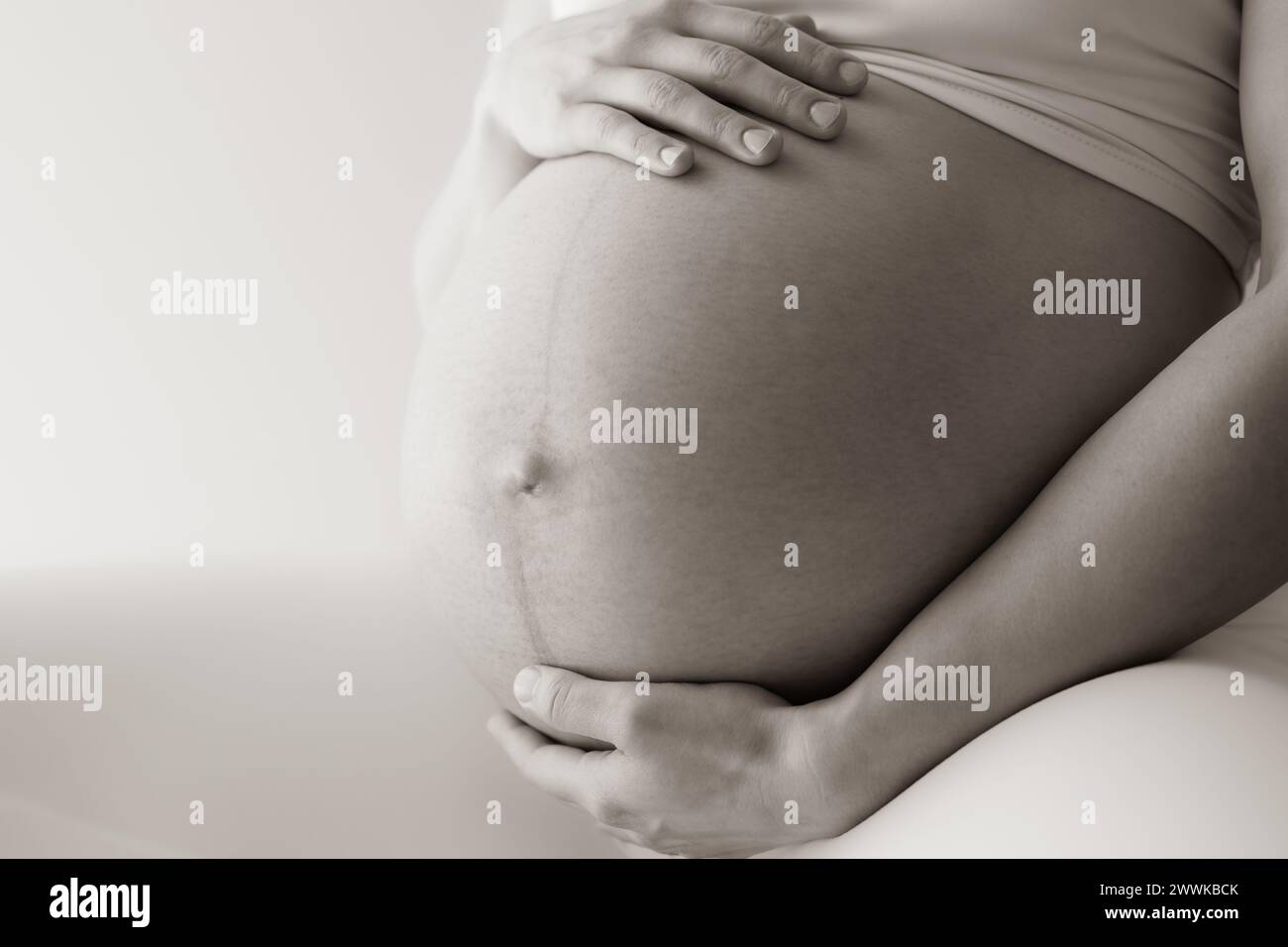 Description: Midsection of a woman sitting and gently holding her very round pregnant baby bump. Side view. White background. Bright shot. Sepia. Stock Photo