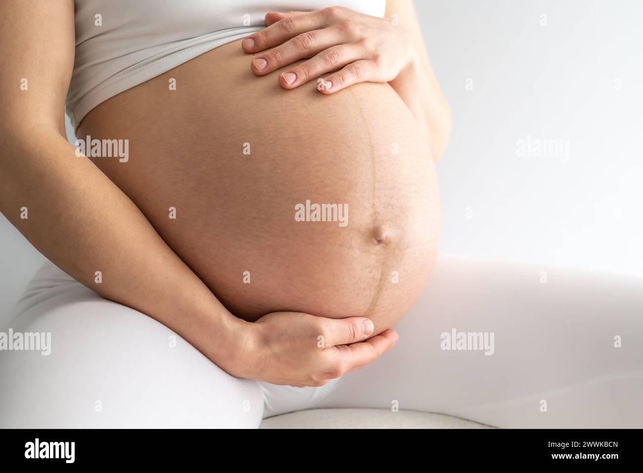 Description: Midsection of a woman sitting on a stool and gently holding her very round pregnant baby belly. Side angle view. White background. Bright Stock Photo