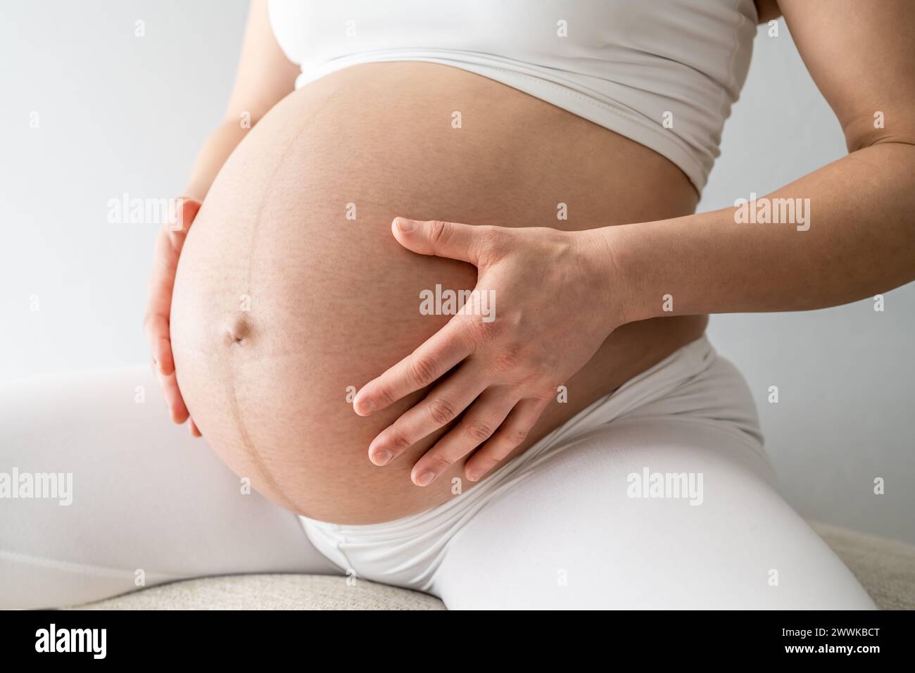 Description: Middle part of a woman sitting on a stool and gently holding her very round pregnant baby belly. Side angle view. White background. Brigh Stock Photo