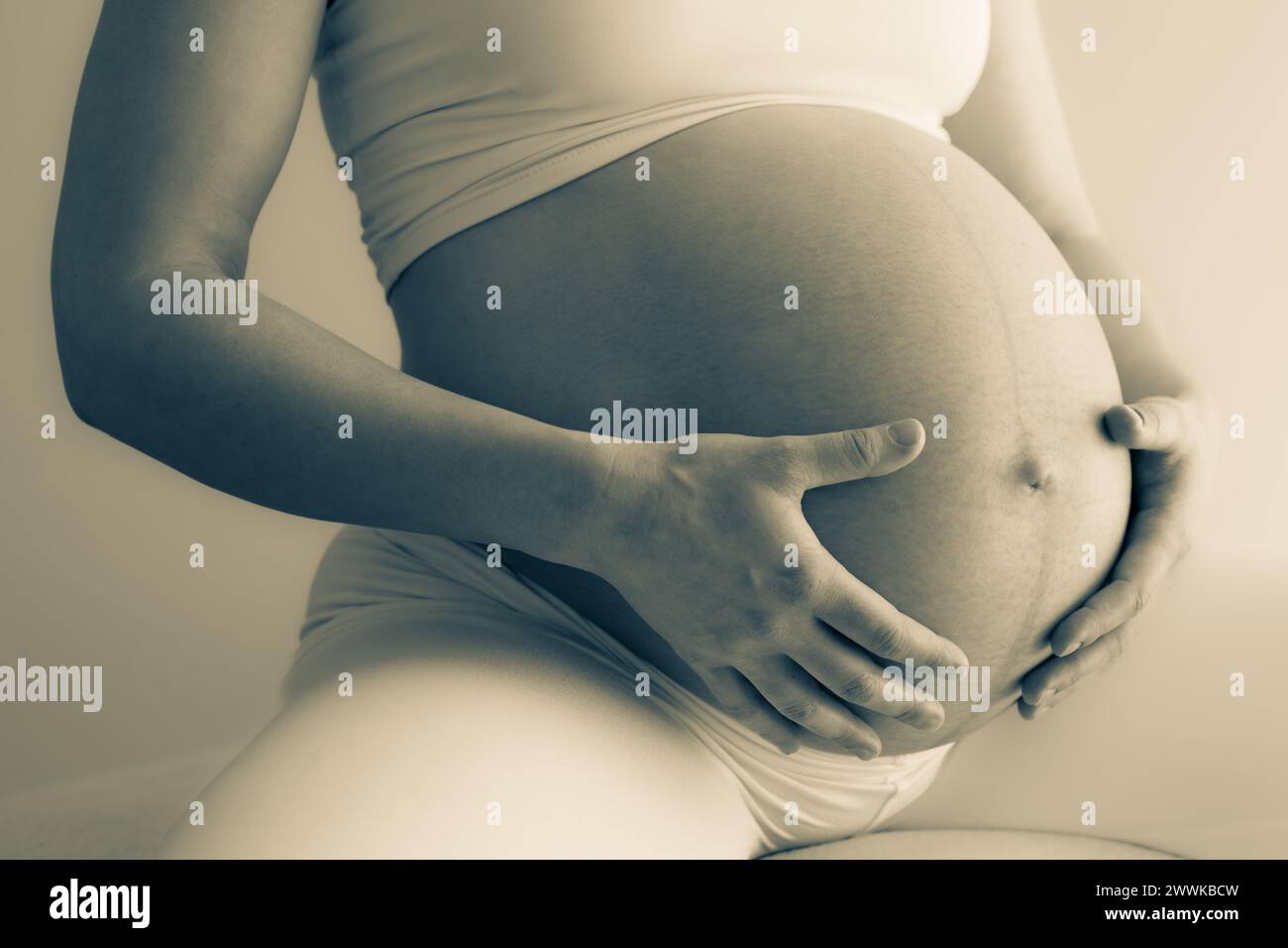 Description: Middle part of a mother sitting on a stool and gently holding her very round pregnant baby belly. Side angle view. White background. Brig Stock Photo
