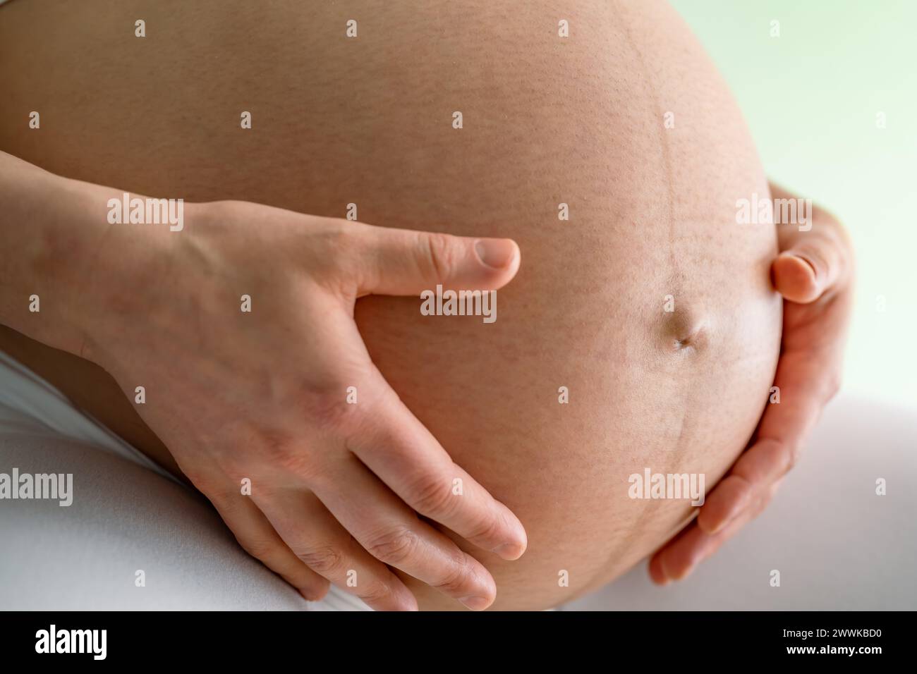 Description: Closeup of a mother sitting and gently holding her very round pregnant baby bump. Side angle view. Green background. Bright shot. Stock Photo