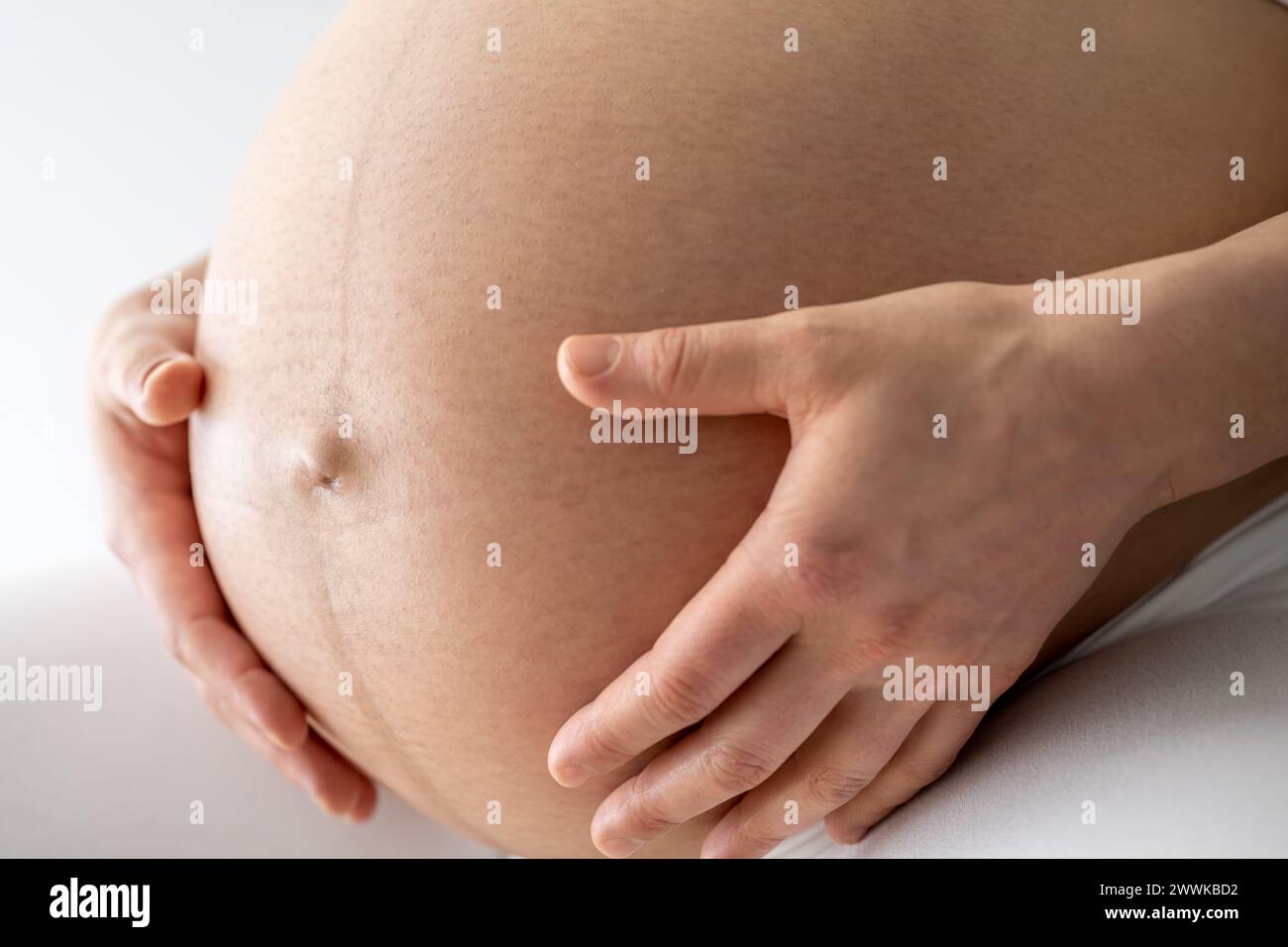 Description: Closeup of a mother sitting and gently holding her very round pregnant baby belly. Side angle view. White background. Bright shot. Stock Photo