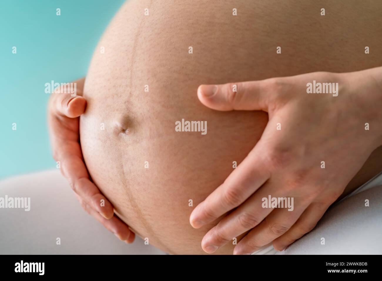 Description: Closeup of a sitting woman gently holding her very round pregnant baby bump. Side angle view. Turquoise background. Bright shot. Black an Stock Photo