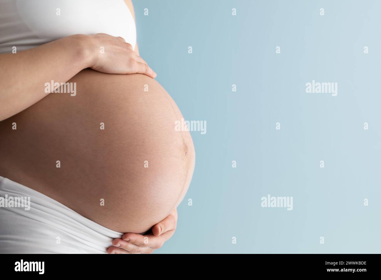Description: Middle part of unrecognizable standing woman in white cloths gently holding her very round pregnant baby bump. Side angle  view. Blue bac Stock Photo