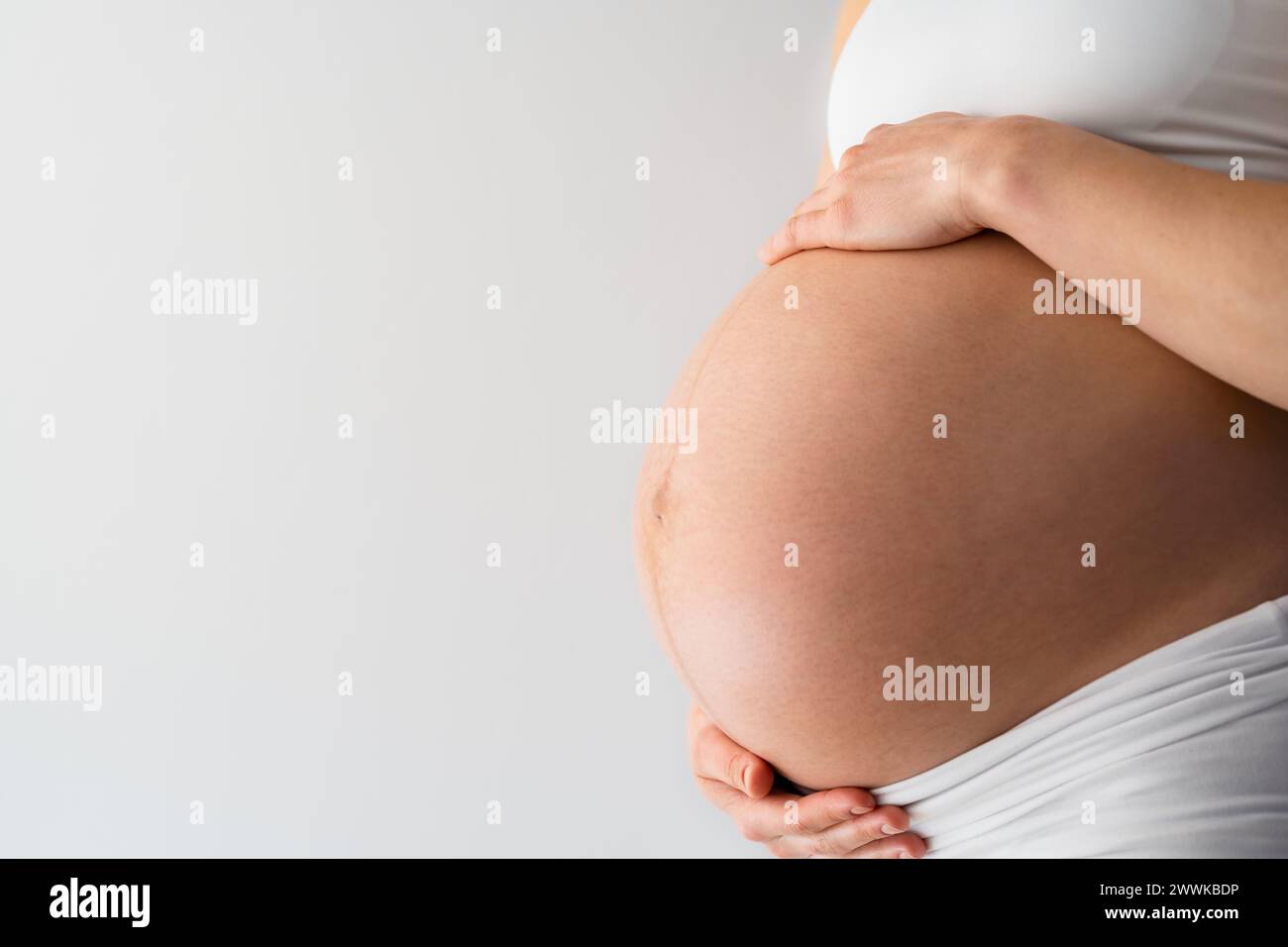 Description: Midsection of unrecognizable standing mother gently holding her very round pregnant baby belly. Side angle  view. White background. Brigh Stock Photo