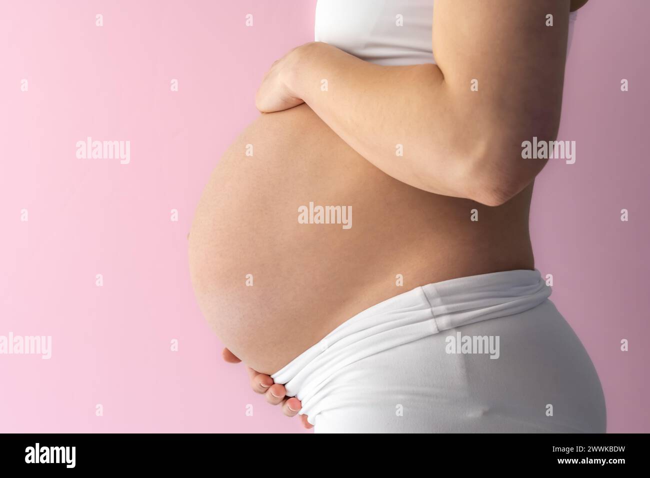 Description: Midsection of unrecognizable standing mother gently holding her very round pregnant baby belly. Side view. Pink background. Bright shot. Stock Photo