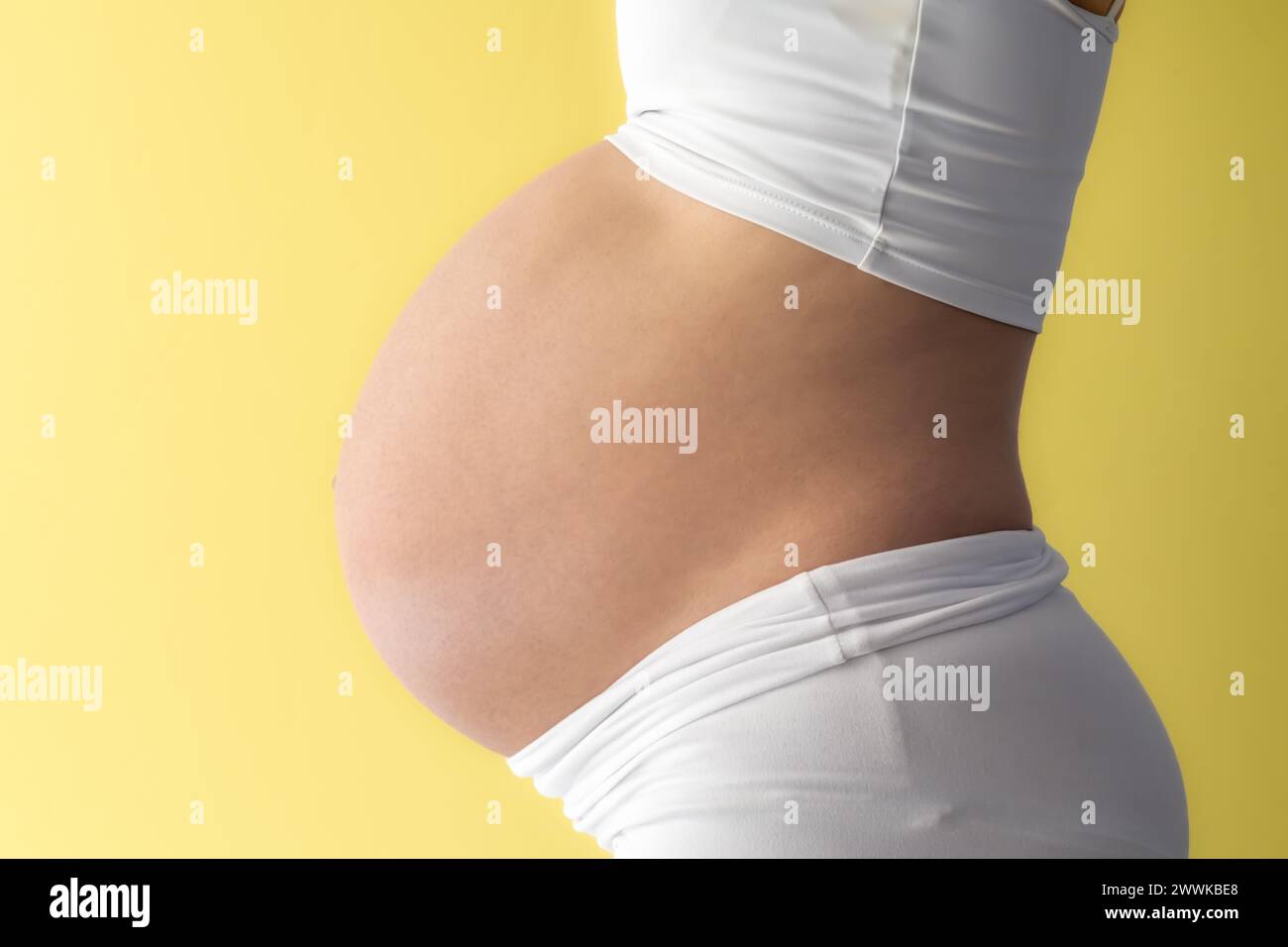 Description: Midsection of unrecognizable standing mother in white cloths with very round pregnant baby belly. Side view. Yellow background. Bright sh Stock Photo