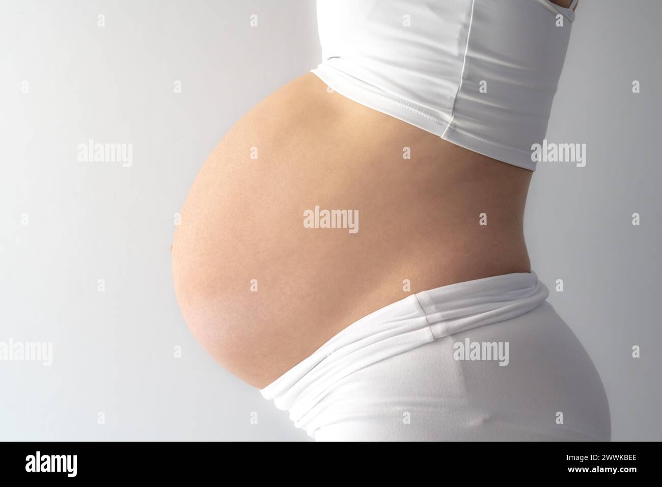 Description: Midsection of unrecognizable standing mother in white cloths with very round pregnant baby belly. Side view. White background. Bright sho Stock Photo